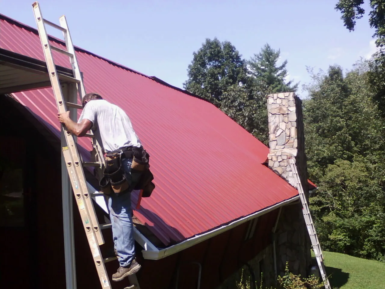 Roofer on a ladder beside a red metal roof and stone chimney — Soffit & Fascia Repair by Roofing Brothers in Chattaroy